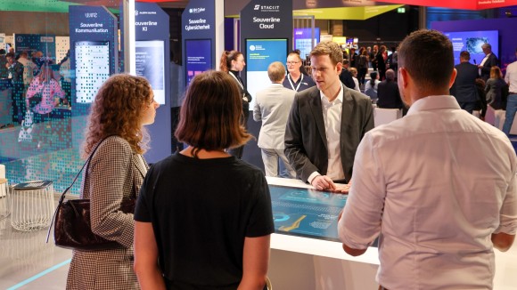 Four people stand at an interactive table at the Smart Country Convention Berlin, in the background stands with topics such as 'Sovereign Communication' and 'Sovereign Cloud'
