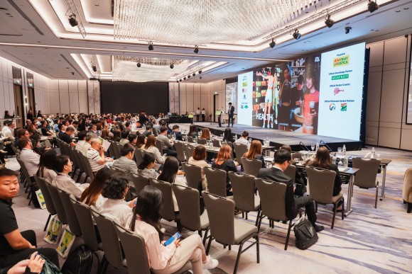 A room full of people sitting on chairs and watching someone present on stage. 