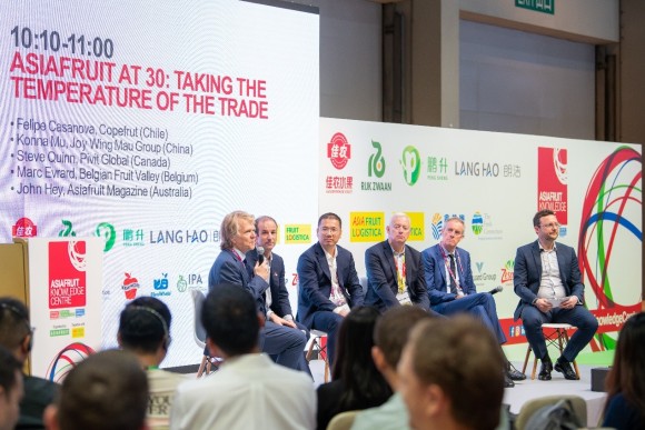 Five men sit on a stage in front of a wall with logos and a screen displaying red text about temperature trade at Asiafruit.