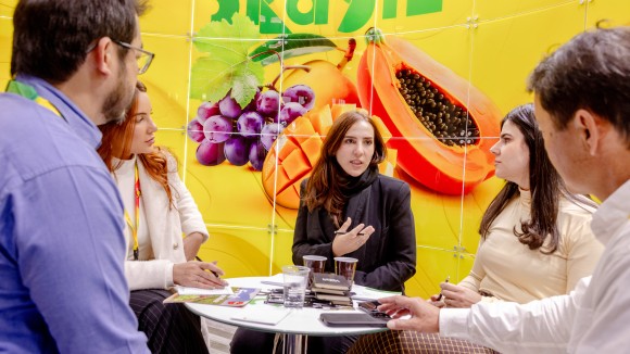 Cinco personas están sentadas alrededor de una mesa conversando. Al fondo se ve un gran mural con frutas.