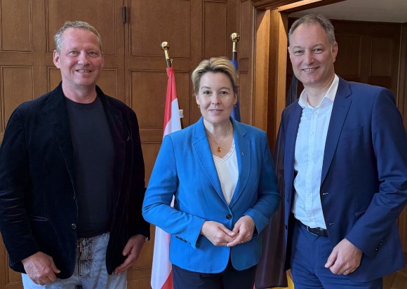 Drei Personen stehen in einem Raum und schauen in die Kamera.