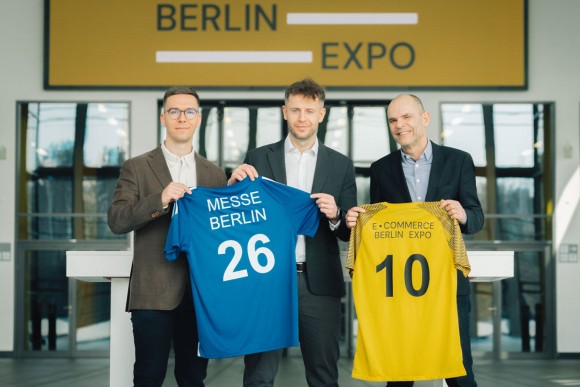 The picture shows three men holding two jerseys in the colours yellow and blue. One jersey reads Messe Berlin and the other E Commerce Berlin Expo.