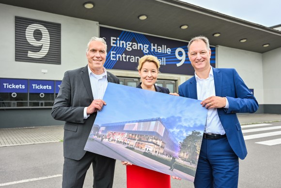 Three people in business attire stand in front of Messe Berlin, holding a rendering of a modern congress hall building.