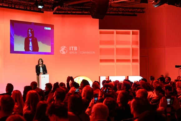 En el escenario de la Convención ITB Berlín 2025, Mitra Sorrells (vicepresidente Senior de Contenido, Phocuswright) se encuentra en un atril frente a una gran audiencia. Detrás de ella, una pared del escenario está iluminada en naranja, s
