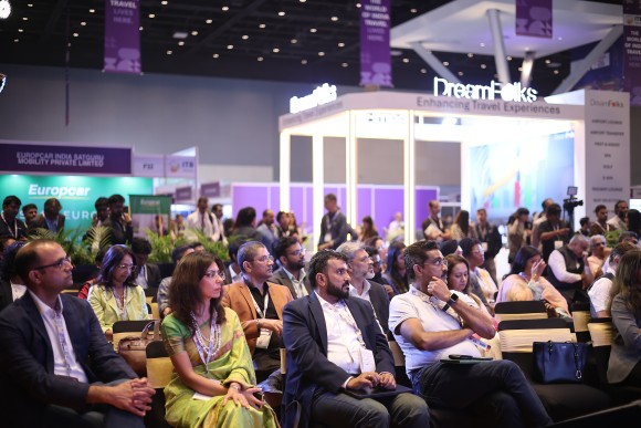 Audience listening attentively to a session, with a trade fair stand in the background.